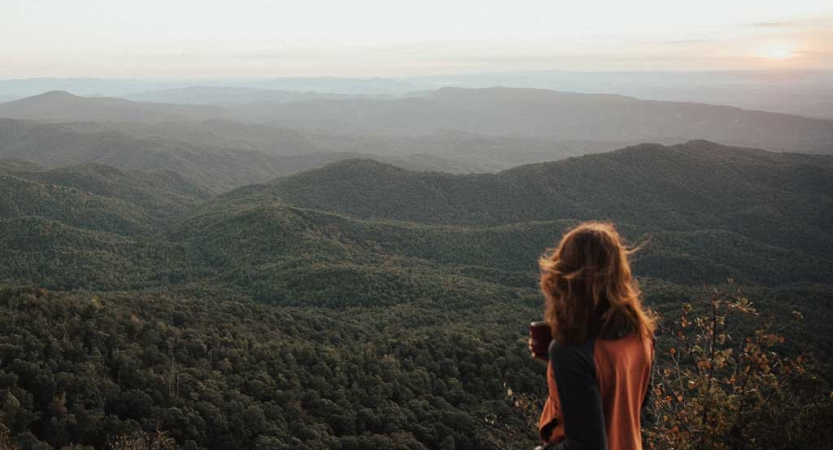 A person with their back to the camera stands on an overlook holding a mug. Below them, the blue ridge mountains stretch for miles. 